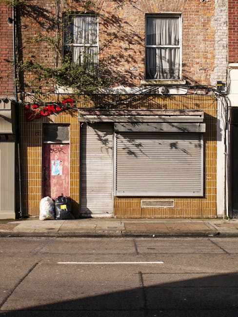 The image depicts the front of a small commercial or residential building with a brick facade, featuring two vertically aligned windows with white frames and curtains on the upper floor. Below, there is a closed, grey metal roll-up shutter covering what appears to be a shop or service entrance, set within a façade with brownish-yellow tiles surrounding it. To the left of the shutter, a slightly narrower grey pedestrian door is also shut. In front of the building, on the pavement, two black garbage bags and a clear plastic bag filled with waste are placed near the door, indicating waste collection or disposal activity. Shadows from nearby trees cast onto the building's facade and pavement, with some greenery visible in the form of a small vine climbing the brick wall near the upper window. The overall scene suggests an urban setting where occasional rubbish removal or on-site waste management services might be employed, consistent with private waste collection practices prevalent in such environments.