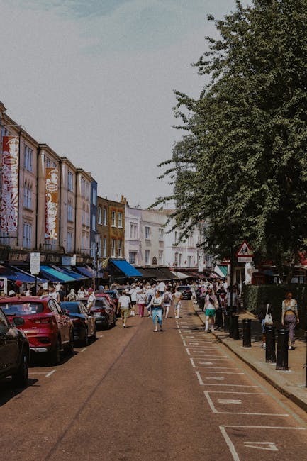 A busy urban street scene with parked cars lining both sides of the road, where a variety of small shops and market stalls with striped awnings are visible on the left. Pedestrians are walking along the pavement and in the street, some carrying bags. The buildings are multi-storey, with a mixture of colorful facades and large windows. On the right side, a large leafy tree provides some shade over the walkway, and there are black bollards separating the pavement from the street. The scene appears to be during daytime with clear lighting. In the background, more shops and pedestrians can be seen, suggesting a bustling marketplace environment, which could involve local or independent waste collection or rubbish removal services such as those offered by Shepherds Bush waste removal providers, in fitting with a comprehensive approach to managing rubbish from busy market areas.
